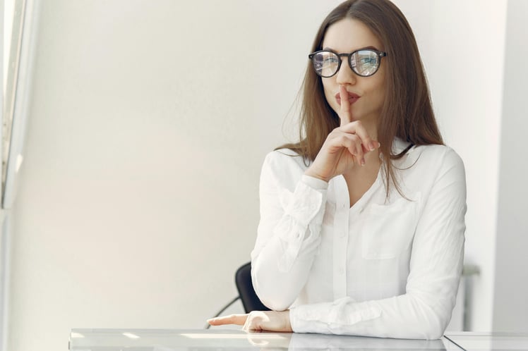 Woman raising her finger to her mouth asking to be quiet while sitting in an office.