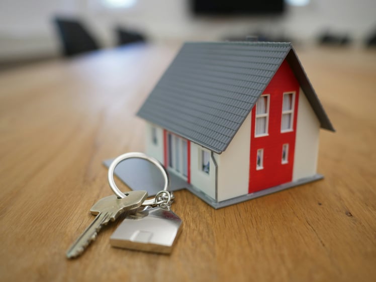 A small model house with a red and white exterior sits on a wooden table next to two keys on a keyring.