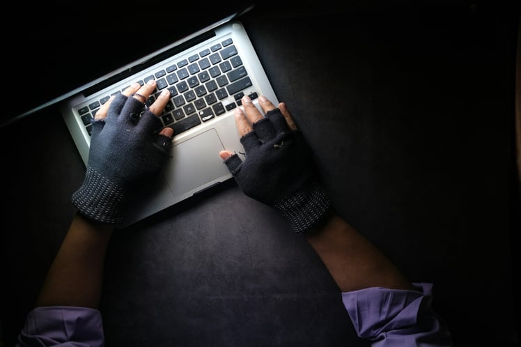 A person wearing fingerless gloves types on a laptop keyboard, illuminated by a light source from above in a dimly lit setting, exuding an aura of security diligence.