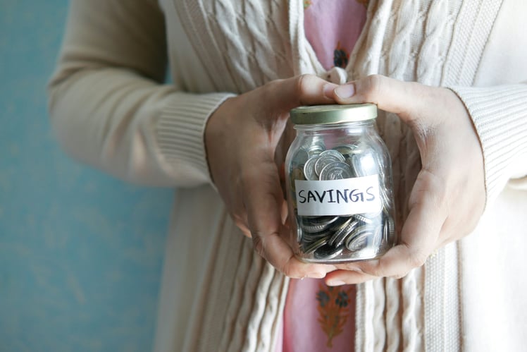A person in a beige sweater holds a glass jar labeled "SAVINGS" filled with coins, planning meticulously for retirement.